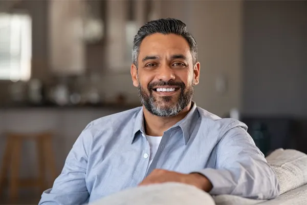 BioTE-Testosterone-Pellet-Clinic A mature South Asian man seated comfortably on a couch, smiling and looking directly at the camera satisfied with BioTE Testosterone Pellet Therapy offered by Scott Gulinson, MD, FACOG in Phoenix.