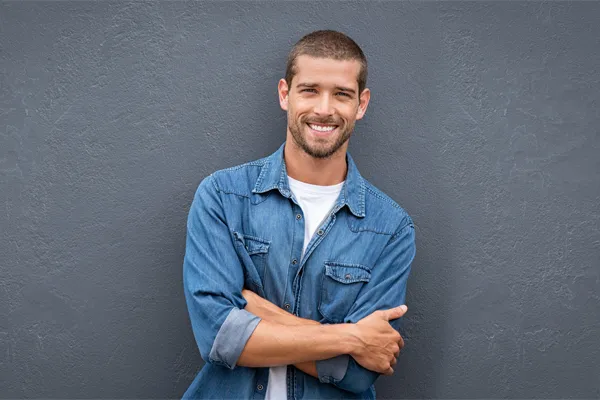 Hormones-for-Men-Doctor A man in a denim shirt stands smiling against a gray-blue wall, pleased with his testosterone hormone therapy from Scott Gulinson, MD, FACOG in Phoenix.