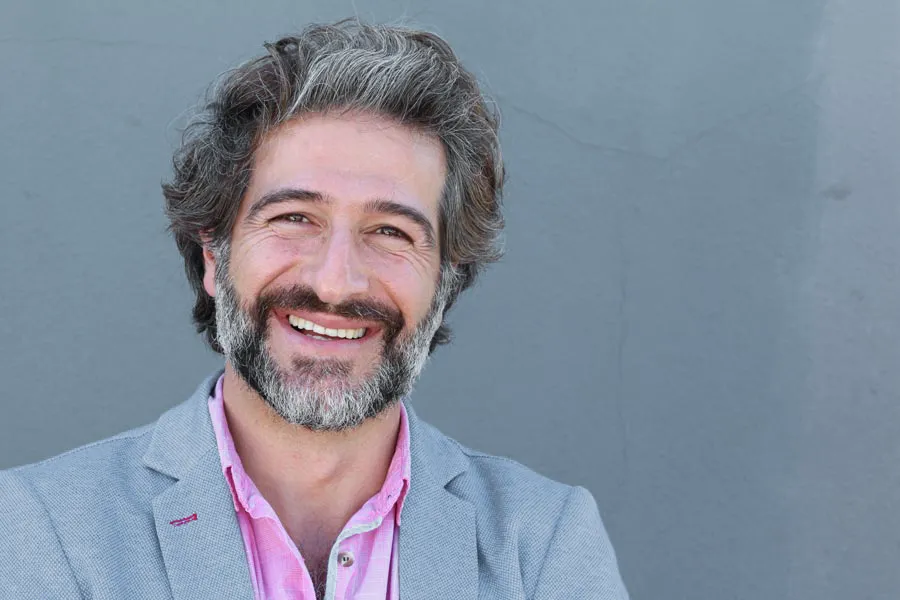 Hypogonadism-Clinic A smiling man with salt and pepper hair and beard wearing a pink shirt and gray blazer stands agains a gray wall. Get hypogonadism treatment from Scott Gulinson, MD, FACOG in Phoenix.