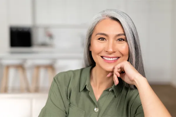 Menopause-Clinic An asian woman with long gray hair and a green shirt sitting and smiling on her couch, happy with her menopause care from [NAME} in Phoenix.